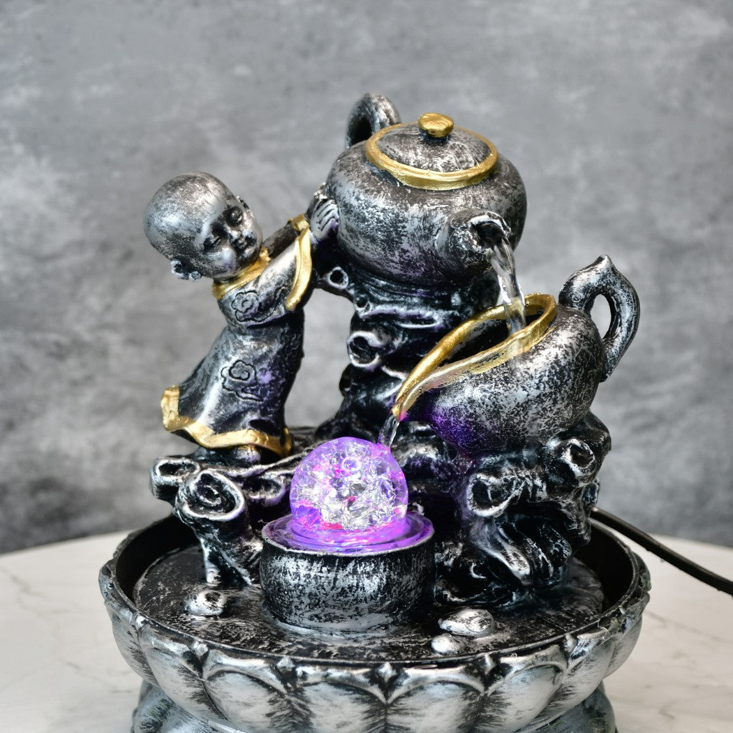 Decorative resin figurines styled as a silver and gold teapot and cup with a small child figure by a tabletop water fountain